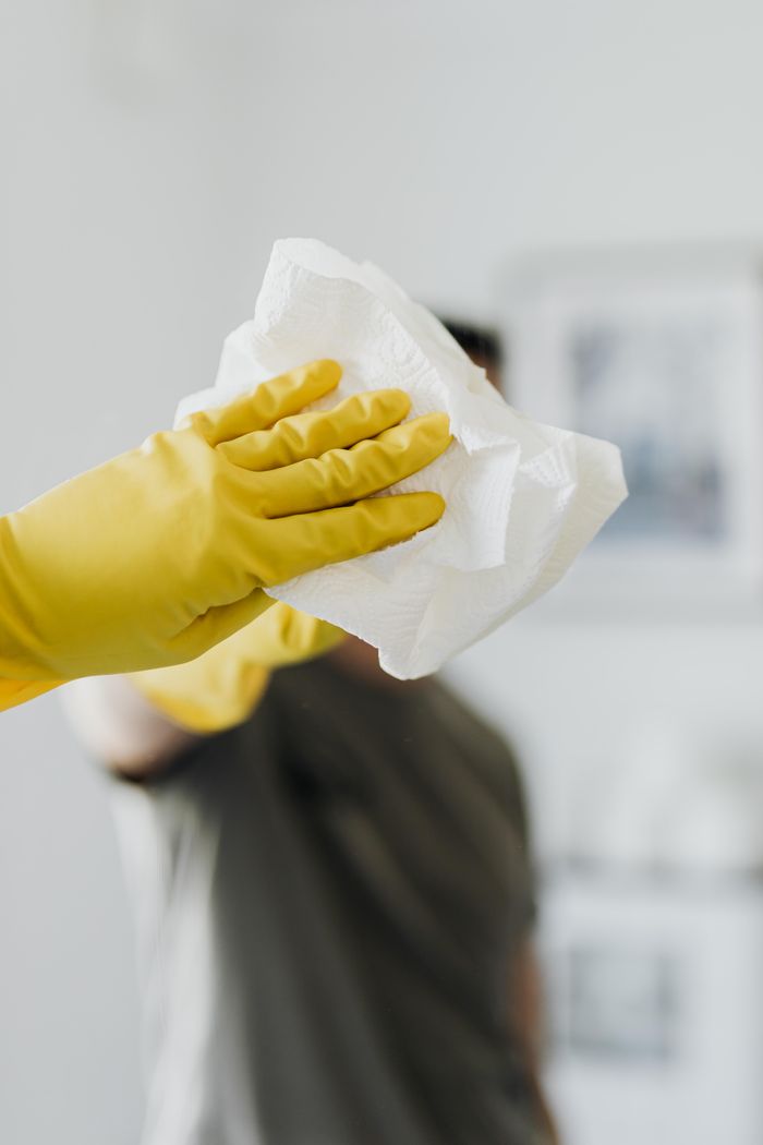Un homme anonyme portant des gants en latex jaunes et un t-shirt foncé essuie le miroir de la salle de bain avec une serviette en papier